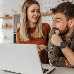 Young couple smiles together as they look at a computer screen in their kitchen.