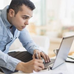 A man sitting on a couch reviews his credit card statement on his laptop.