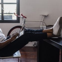 Happy man has his feet up on his desk as he leans back in his chair and looks at his laptop in his lap.