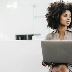 Woman looking off into the distance sits with laptop on her knees.