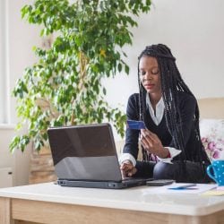 Young woman sits on her couch working on a laptop and holding a credit card in her hand.