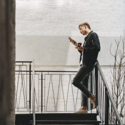 A young man stands in stairwell holding paper coffee cup and scans his mobile phone.