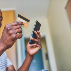 A man looks down at a credit card held in one hand and mobile phone in the other hand.
