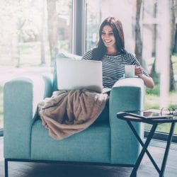 Smiling young woman, her legs covered with a blanket, sits in a chair looking at her laptop and holding a cup of coffee.