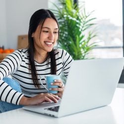 Happy young woman sitting at her table, focused on her laptop, and enjoying a cup of coffee.