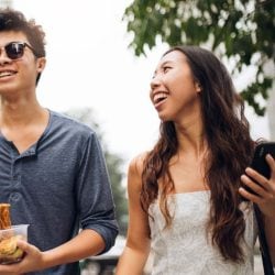 Two friends walking outside and smiling, one holding a phone and one eating.