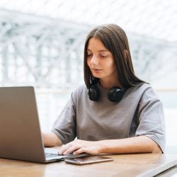 A dark-haired woman is sitting at a table while browsing on a laptop.