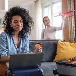 Woman uses a credit card while looking at her laptop as her teenage daughter plays with a toy airplane behind her.