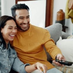 A smiling couple sit closely together on a couch. The man holds a TV remote.