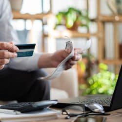 A person holds a credit card and some papers in their hand as they sit in front of a laptop.