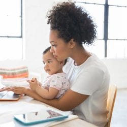With a toddler in her lap, a young woman sits at a kitchen table and works on her laptop.