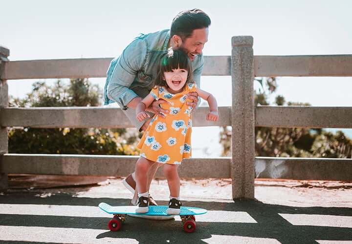 Father and daughter laughing as he pushes her on a skateboard.
