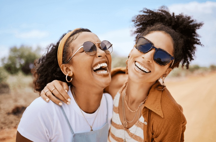 Two women wearing sunglasses standing outside laughing.