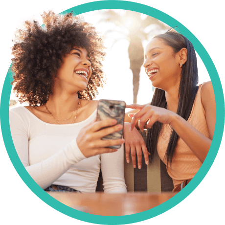 Two women sitting outside smiling at each other while using mobile device.