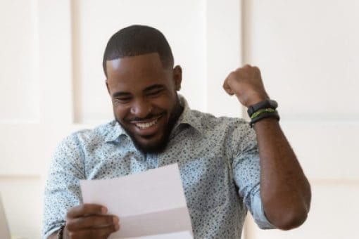 Man sitting at a desk in front of laptop smiles and pumps his fist as he reads from a folded piece of paper.