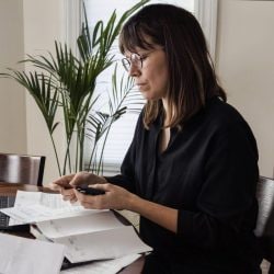 A woman sits at her kitchen table with her laptop and reviews her paper credit card statements.
