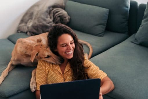 A young lady sitting near the sofa with her laptop laughs as her dog hugs her from behind