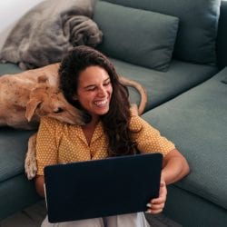 A young woman sitting on the floor and holding a laptop laughs as a dog nuzzles her neck.