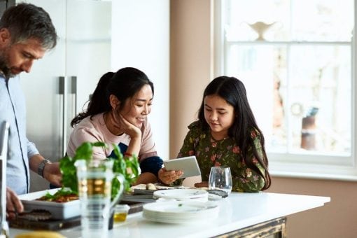 Daughter shows mom digital tablet while husband prepares dinner.