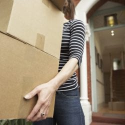 Woman carrying cardboard boxes in front of an open door to a house.