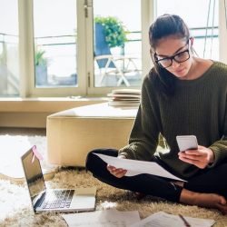 Young woman sits cross-legged on floor holding mobile phone and papers.