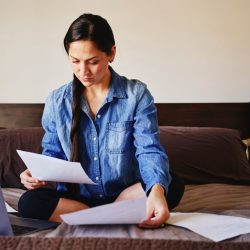 A woman in a blue shirt sits on her bed looking at a paper bill.