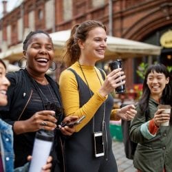 Young women walk down the street laughing and drinking coffee.