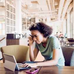 Young woman sits on a coffee shop chair working on a laptop.