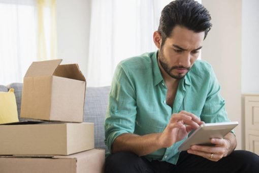 A man uses a tablet as he sits on a couch next to a pile of cardboard boxes.