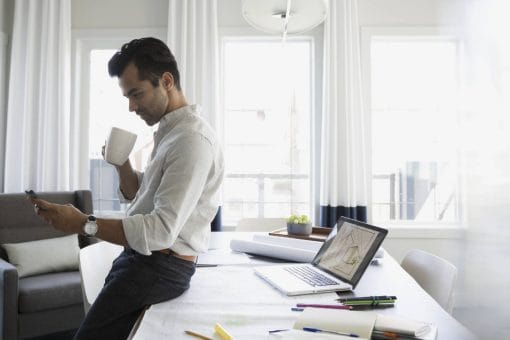 Professional man drinks coffee and leans against his desk while looking at his mobile phone