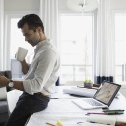 A young man leans against his desk and looks at his phone.