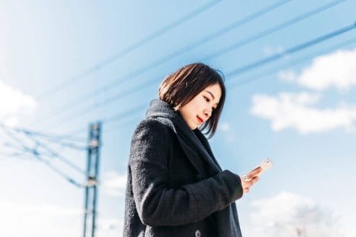 Woman in winter coat stands outside looking at mobile phone in her hand.