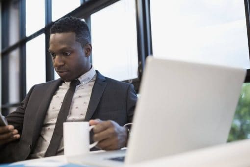 Businessman sits at desk holding coffee mug and looking at mobile phone.