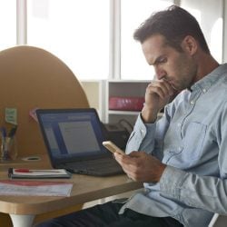 Man sits at a desk in his office, looking down at his cell phone.