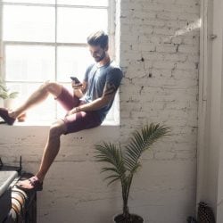 Young man sits on a windowsill in his apartment looking down at his mobile phone.