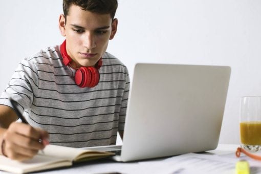 Young man with red headphones around his neck writes in notebook.