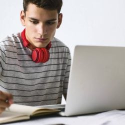 Young man in a black striped shirt wears red headphones around his neck while writing in a notebook.