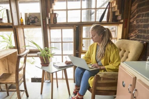 A woman sits in a sunroom writing in a notebook with a laptop on her lap.