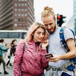 A happy traveling couple on a busy street look at the man’s mobile phone