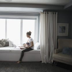 Mom with infant sits on windowsill looking at a laptop.