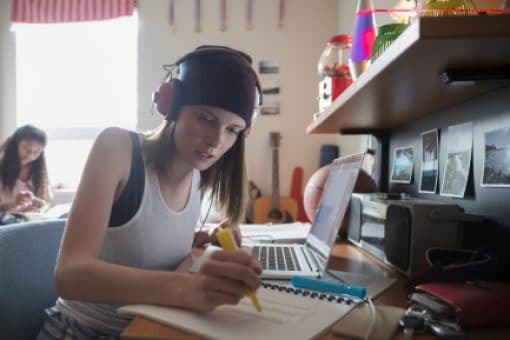 Young female student wearing headphones sits at her desk highlighting a notebook