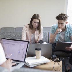 College students sit together in a common room while working on laptops and a tablet.
