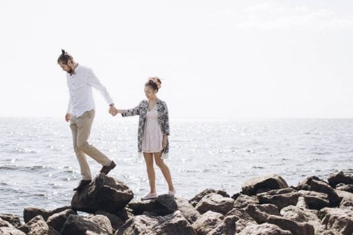 A couple walks on rocks near the ocean.