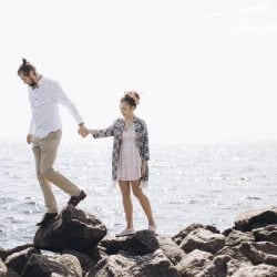 A couple walks on rocks near the ocean while traveling.