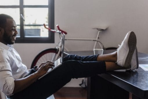 Happy man has his feet up on his desk as he leans back in his chair and looks at his laptop in his lap