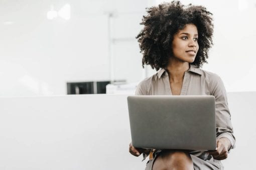 Woman looking off into the distance sits with laptop on her knees.