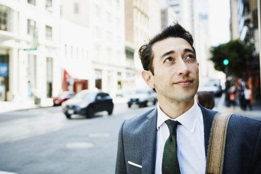 A young professional man looks up toward the sky as he walks down a street.