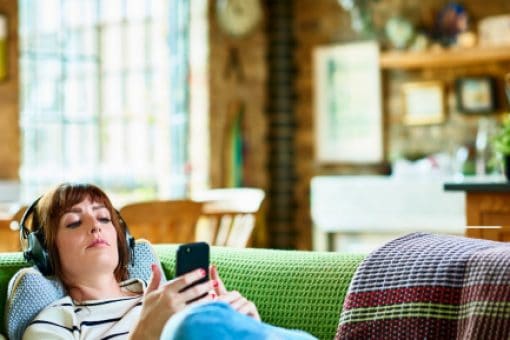 Woman wearing large headphones reclines on couch while looking at mobile phone.