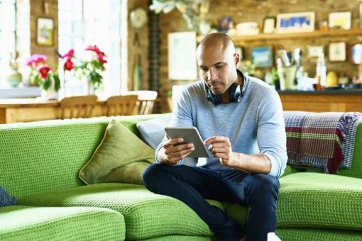 Man with headphones hung around his neck sits on a couch looking at a digital tablet