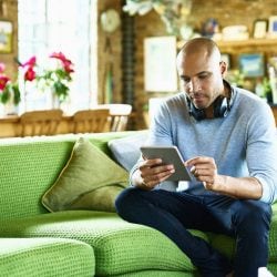 Man with headphones hung around his neck sits on a couch looking at a digital tablet.